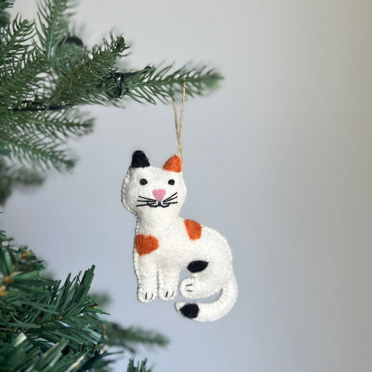 May include: A white felt cat ornament with orange and black spots. The cat has a black nose and pink cheeks. The ornament is hanging from a brown string.