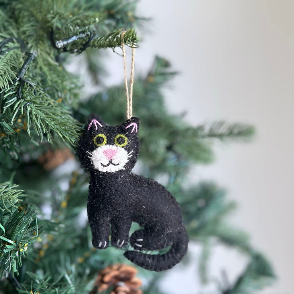May include: Black felt cat ornament with green eyes, pink nose and mouth, hanging from a string on a green Christmas tree.