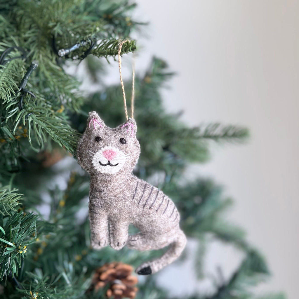 May include: A gray felt cat ornament with pink nose and black eyes hanging on a Christmas tree.