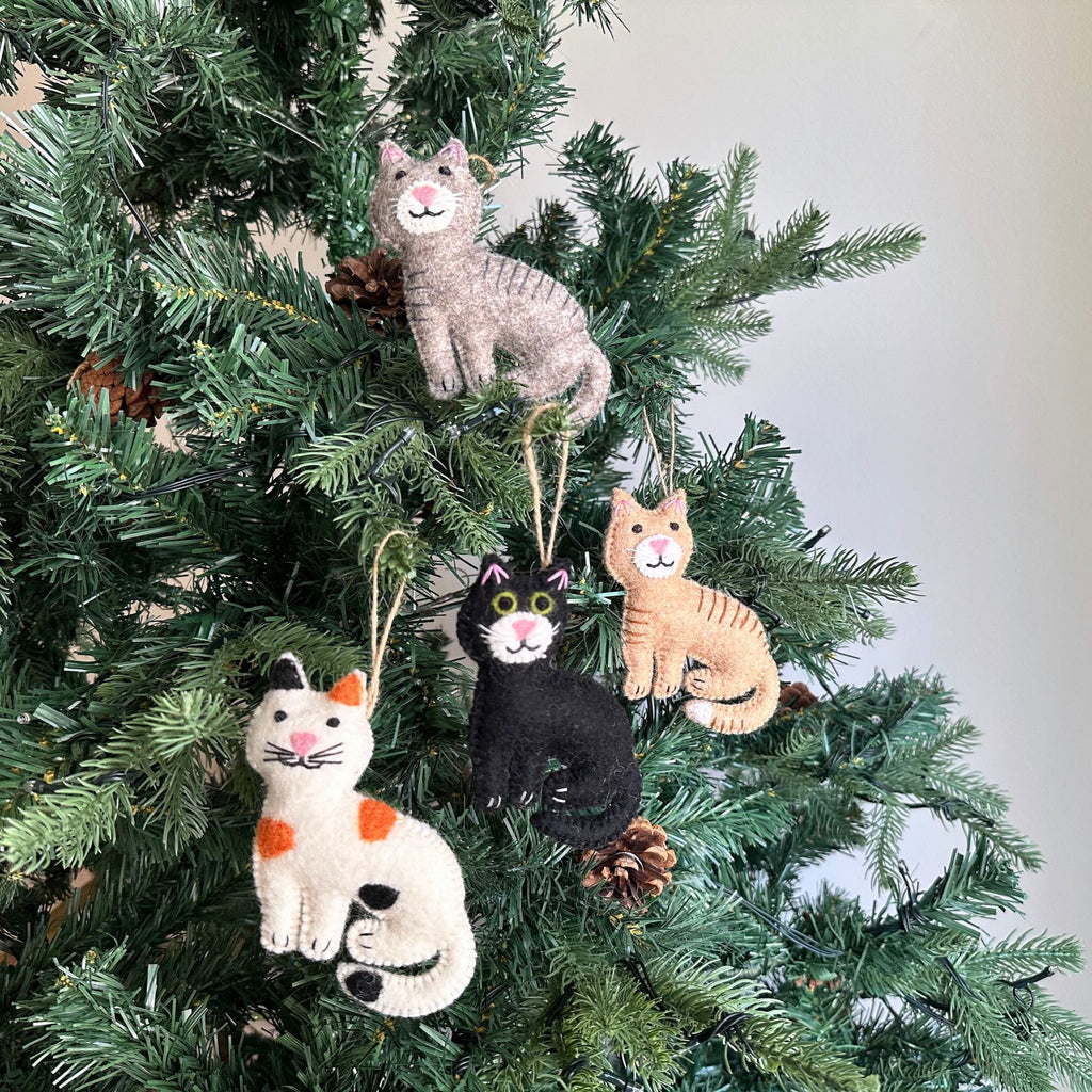 May include: Four felt cat ornaments hanging on a green Christmas tree. The cats are different colors: gray, black, orange, and white with orange spots. Each cat has a white face with black eyes and a pink nose.