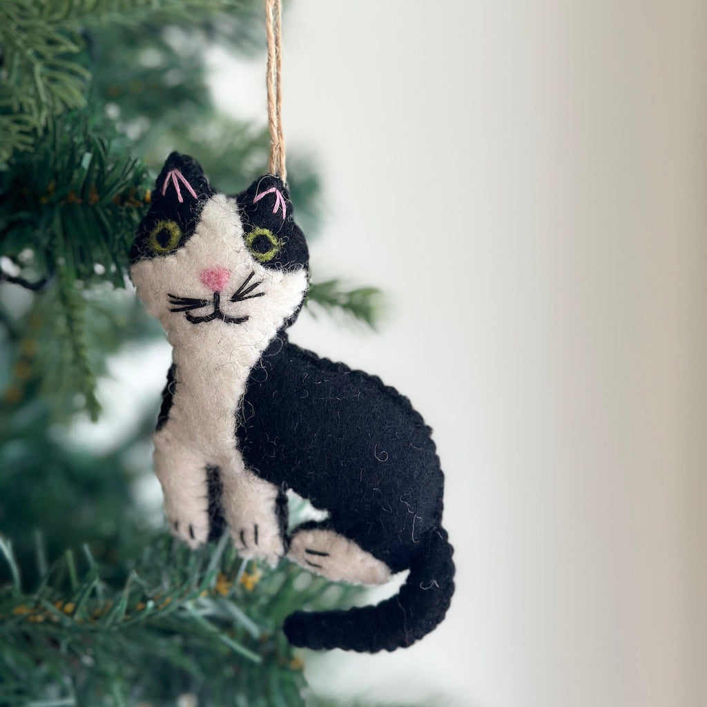 May include: A black and white felt cat ornament with green eyes and pink ears. The cat is sitting on a green Christmas tree branch.