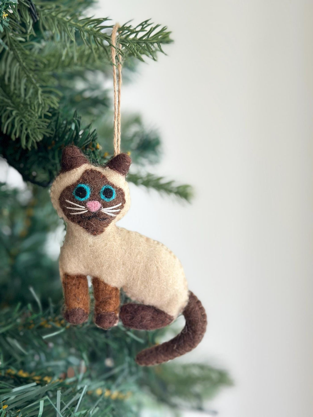 May include: A felt Siamese cat ornament with blue eyes and a brown tail, hanging from a tree branch.