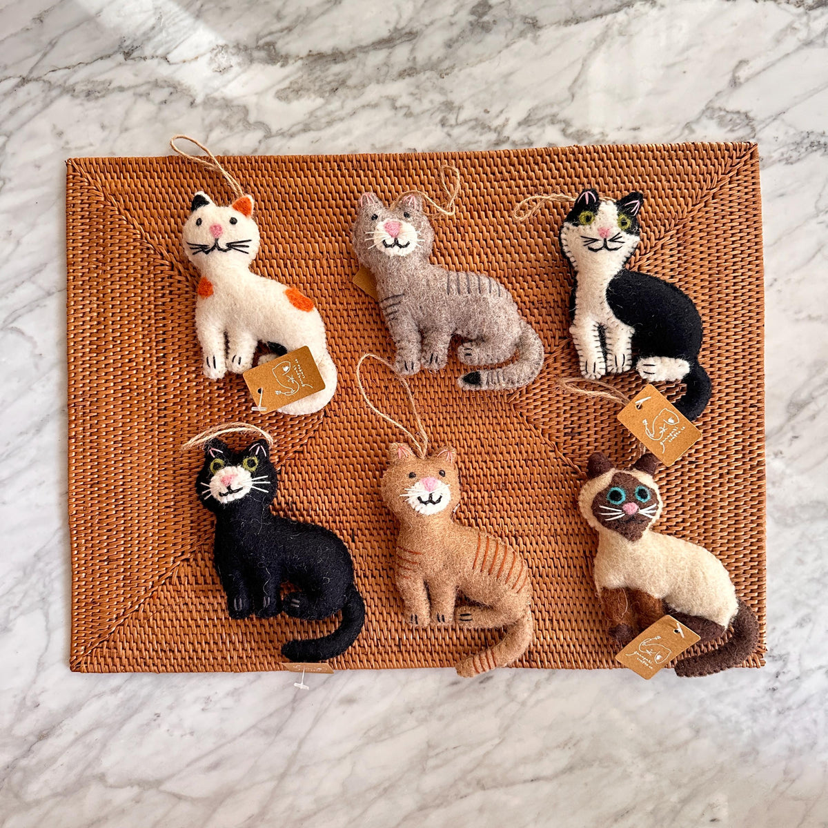 May include: Six felt cat ornaments with different colored fur and facial expressions. The ornaments are hanging from a brown wicker surface.