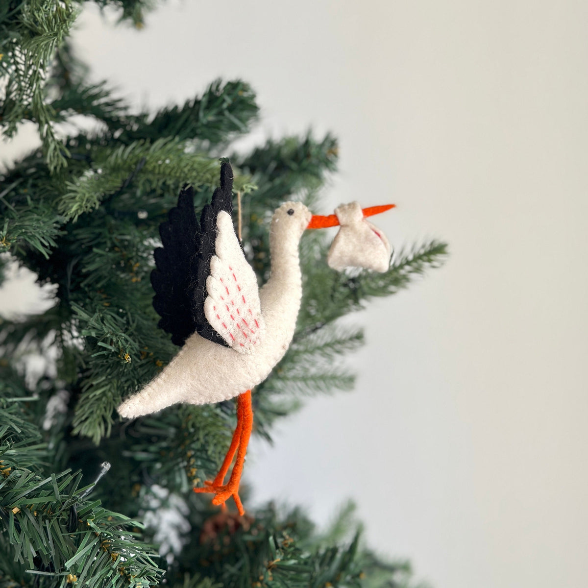 May include: A white felt stork ornament with black wings and orange legs, carrying a small white felt bundle in its beak. The ornament is hanging from a Christmas tree.