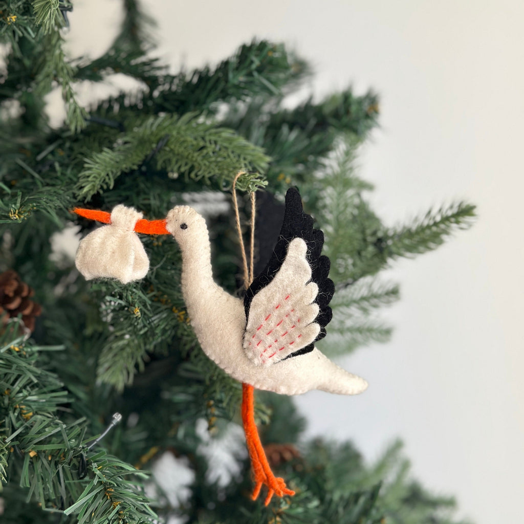 May include: A white felt stork ornament with black and red wings, carrying a white felt bundle in its beak. The ornament is hanging from a tree branch.