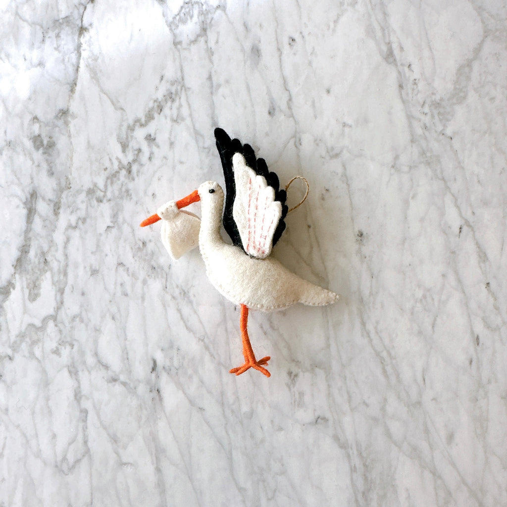 May include: A white felt stork ornament with black and pink wings, holding a white felt bundle in its beak. The stork has orange legs and a black eye.