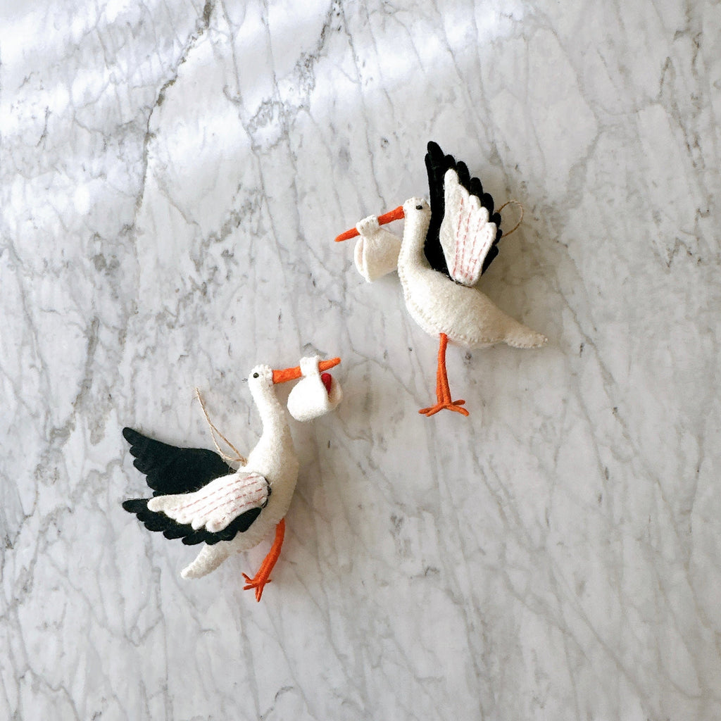 May include: Two white felt stork ornaments with black wings and orange beaks. Each stork is holding a small white felt bundle in its beak.