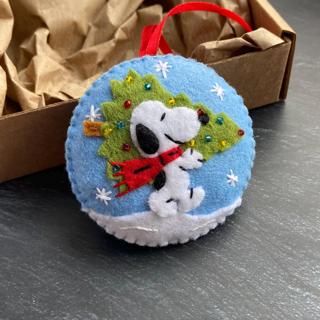 May include: A round, blue felt Christmas ornament featuring Snoopy with a red scarf and a green Christmas tree. White snowflakes and a white snow base surround the character. A red ribbon is attached for hanging. The ornament is inside a brown cardboard box.
