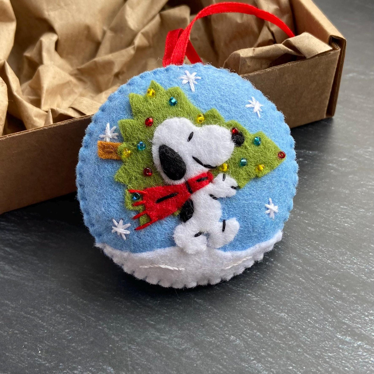 May include: A round, blue felt Christmas ornament featuring Snoopy with a red scarf and a green Christmas tree. White snowflakes and a white snow base surround the character. A red ribbon is attached for hanging. The ornament is inside a brown cardboard box.