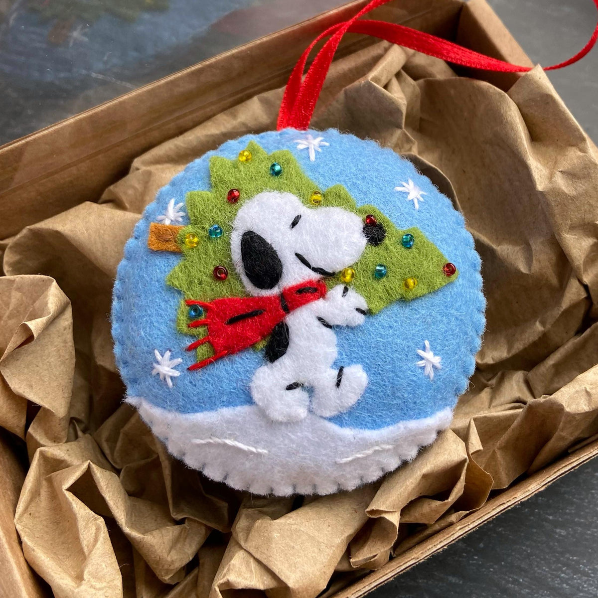 May include: A round, felt Christmas ornament featuring Snoopy wearing a red scarf, standing in front of a green Christmas tree. The ornament is on a blue background with white snowflakes and a white snow base. A red ribbon is attached for hanging.