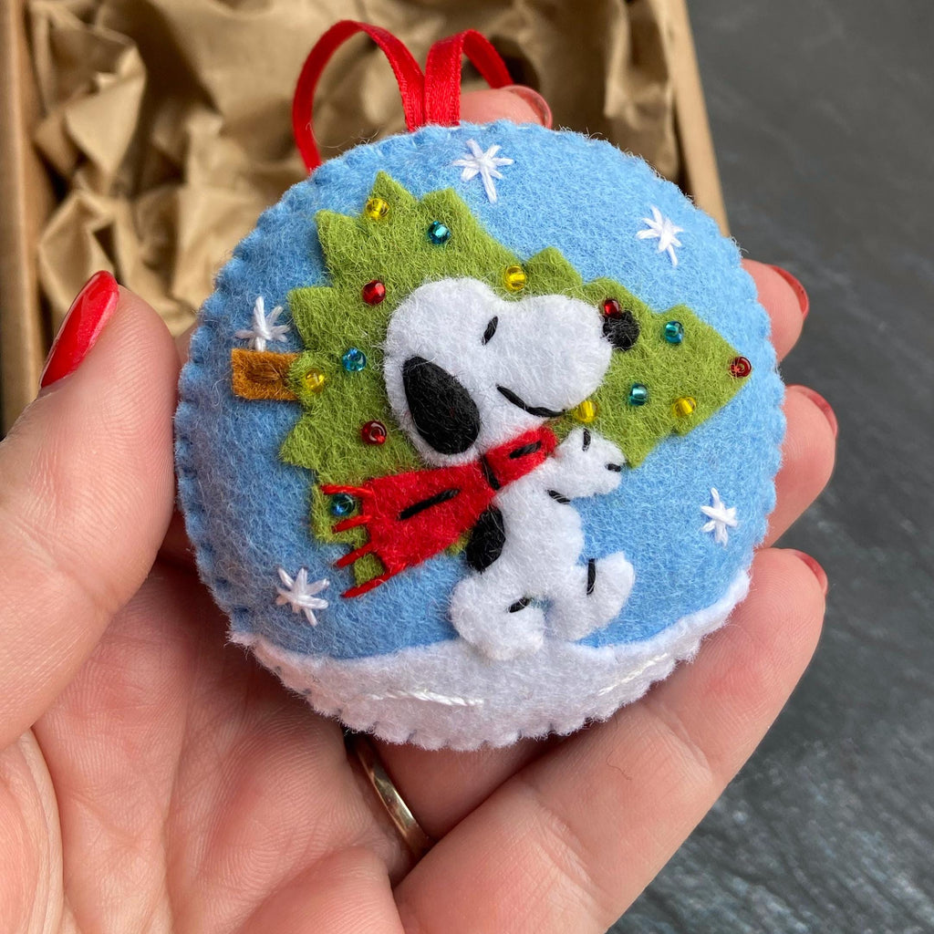 May include: A round, blue felt Christmas ornament featuring Snoopy with a red scarf and a green Christmas tree. White snowflakes and a white snow base decorate the ornament. A red ribbon is attached for hanging.