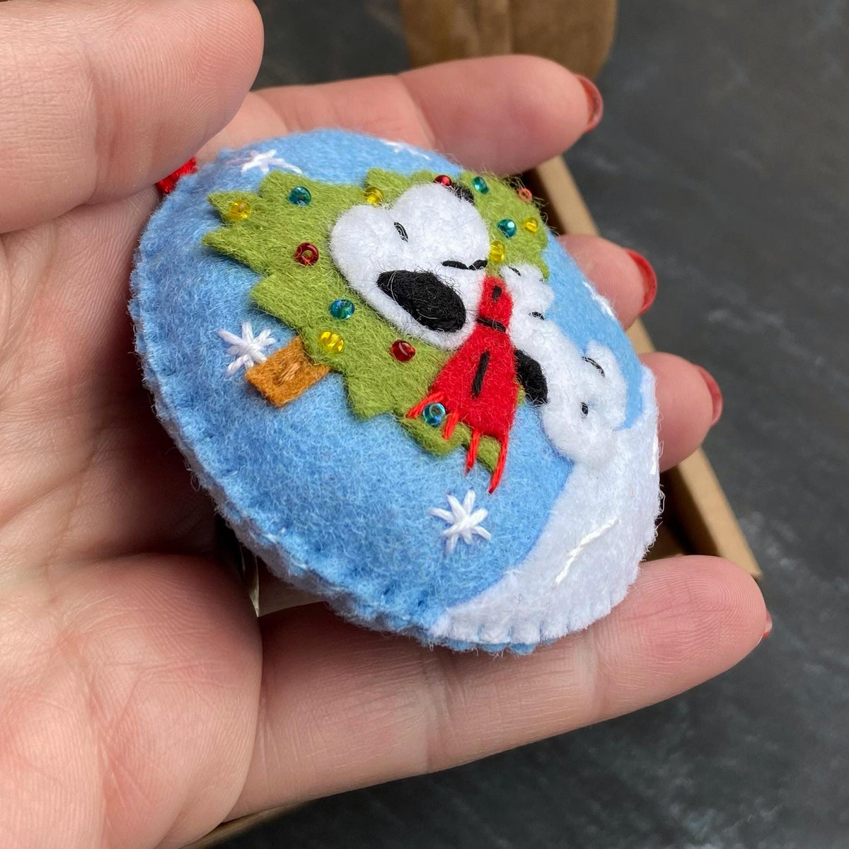 May include: A handmade felt Christmas ornament featuring Snoopy and a Christmas tree on a blue background. The ornament has white snowflakes and a red scarf. The ornament is oval-shaped and has a red loop for hanging.
