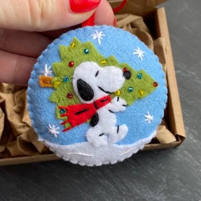 May include: A round, blue felt Christmas ornament featuring Snoopy carrying a green Christmas tree. The ornament has white snowflakes and a white snow base. Snoopy wears a red scarf. A red ribbon is attached for hanging.