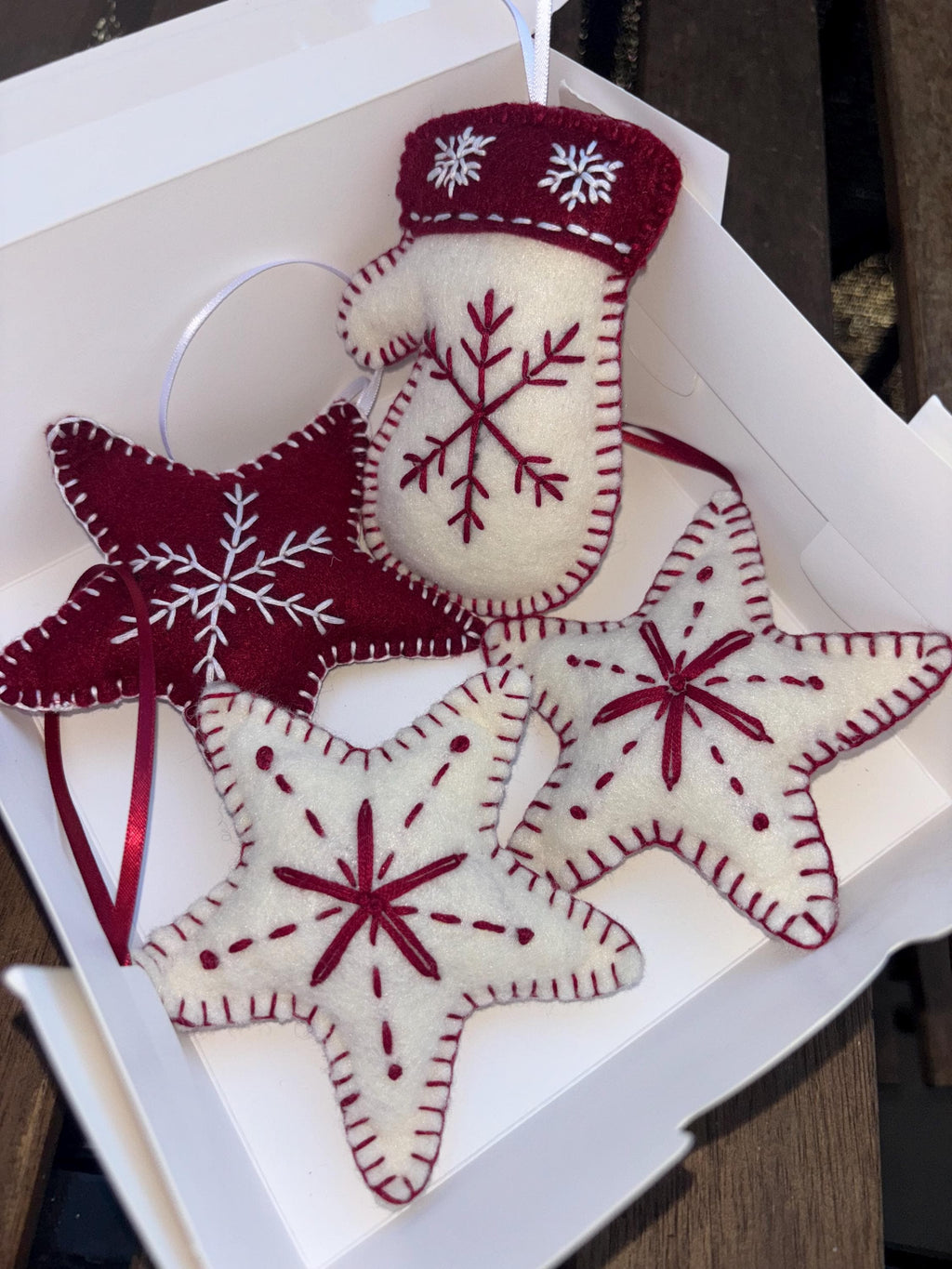 May include: A set of handmade Christmas ornaments in a white box. The ornaments include red and white felt stars and a mitten, all decorated with snowflake designs and stitched details. Each ornament has a ribbon for hanging.