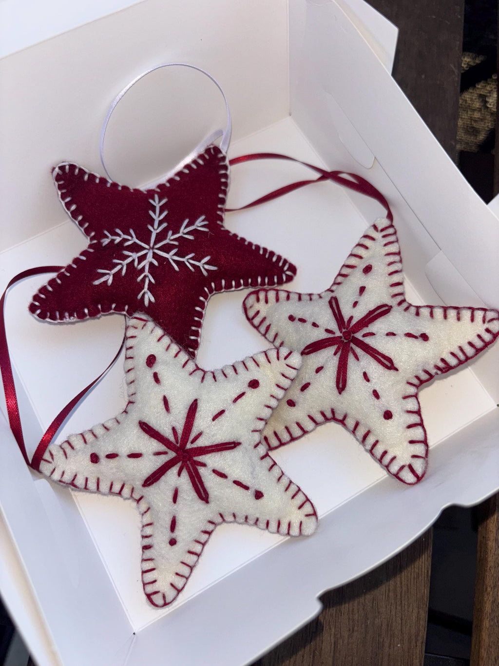 May include: Three star-shaped Christmas ornaments in a white box. Two are off-white with red stitching and starburst designs. The third is burgundy with white snowflake embroidery. Each has a red ribbon for hanging.
