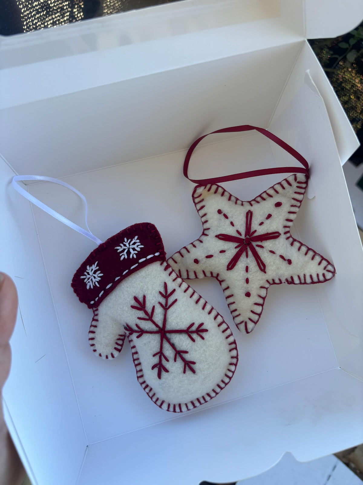 May include: Two felt Christmas ornaments: a mitten and a star. The mitten is white with a burgundy cuff and snowflake embroidery. The star is white with burgundy stitching and a burgundy center. Both have red ribbon loops for hanging.