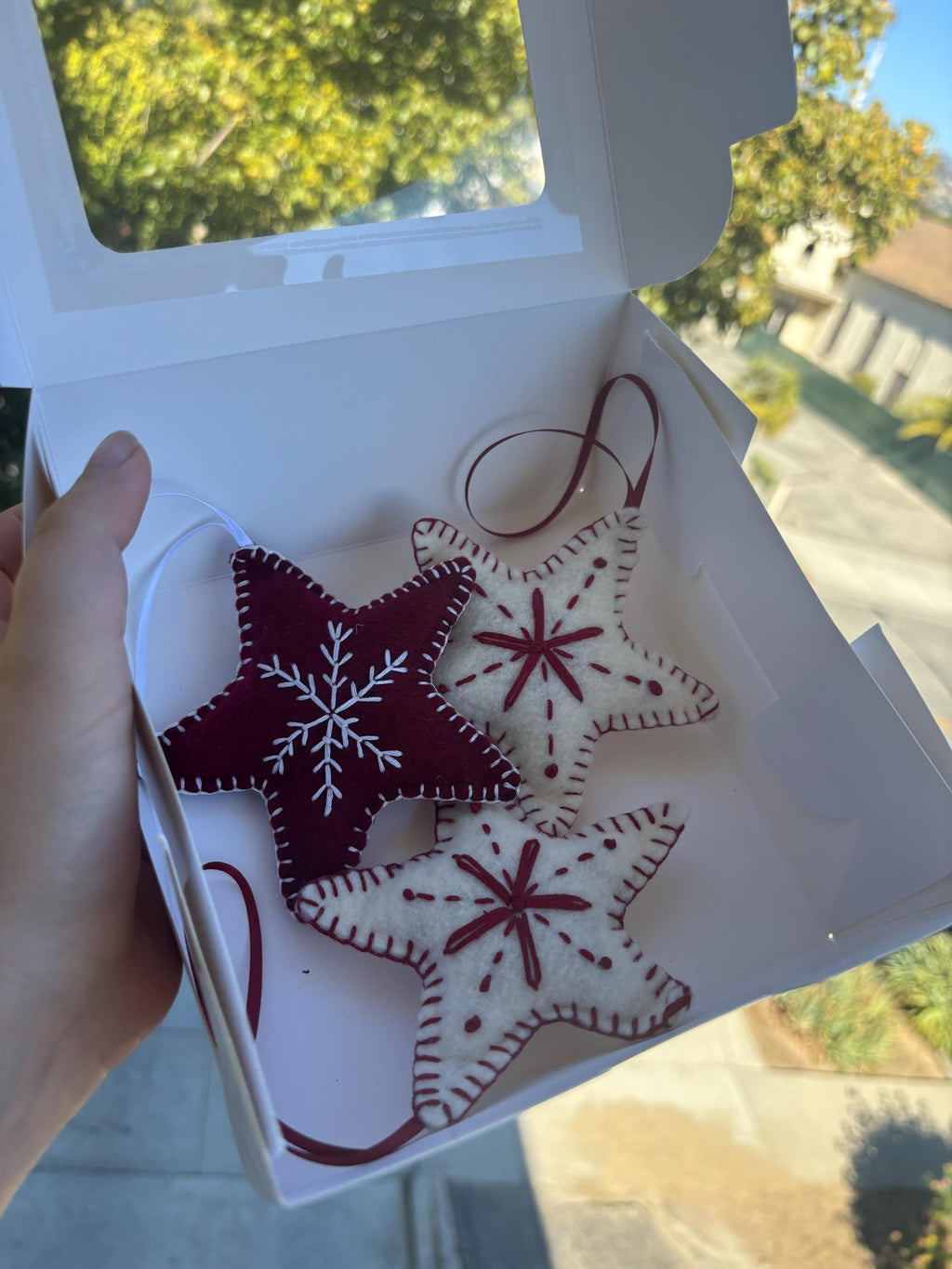May include: A white box holds three star-shaped Christmas ornaments. Two are off-white with red embroidered details, and one is burgundy with a white snowflake design. Each has a loop for hanging.