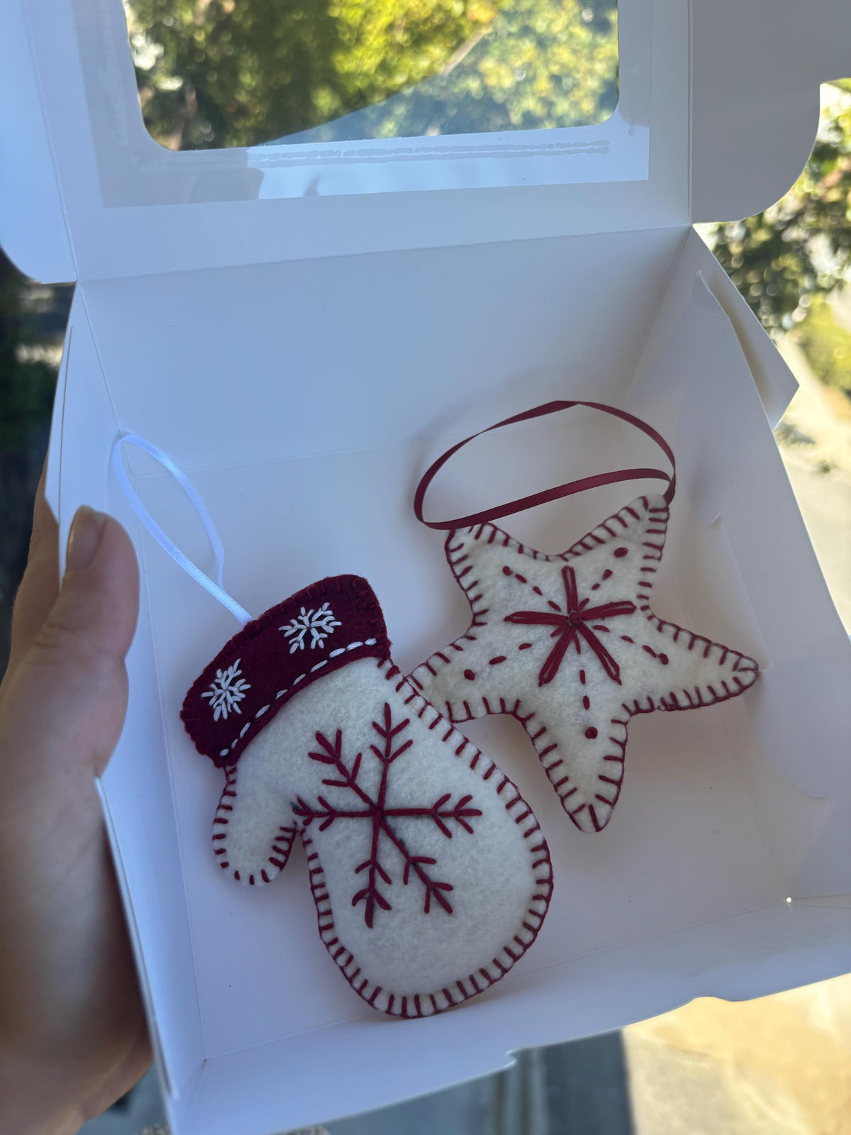 May include: Two handmade Christmas ornaments: a white mitten and a star. The mitten has a burgundy cuff and snowflake details. The star is white with burgundy stitching and a burgundy center. Both have hanging ribbons.
