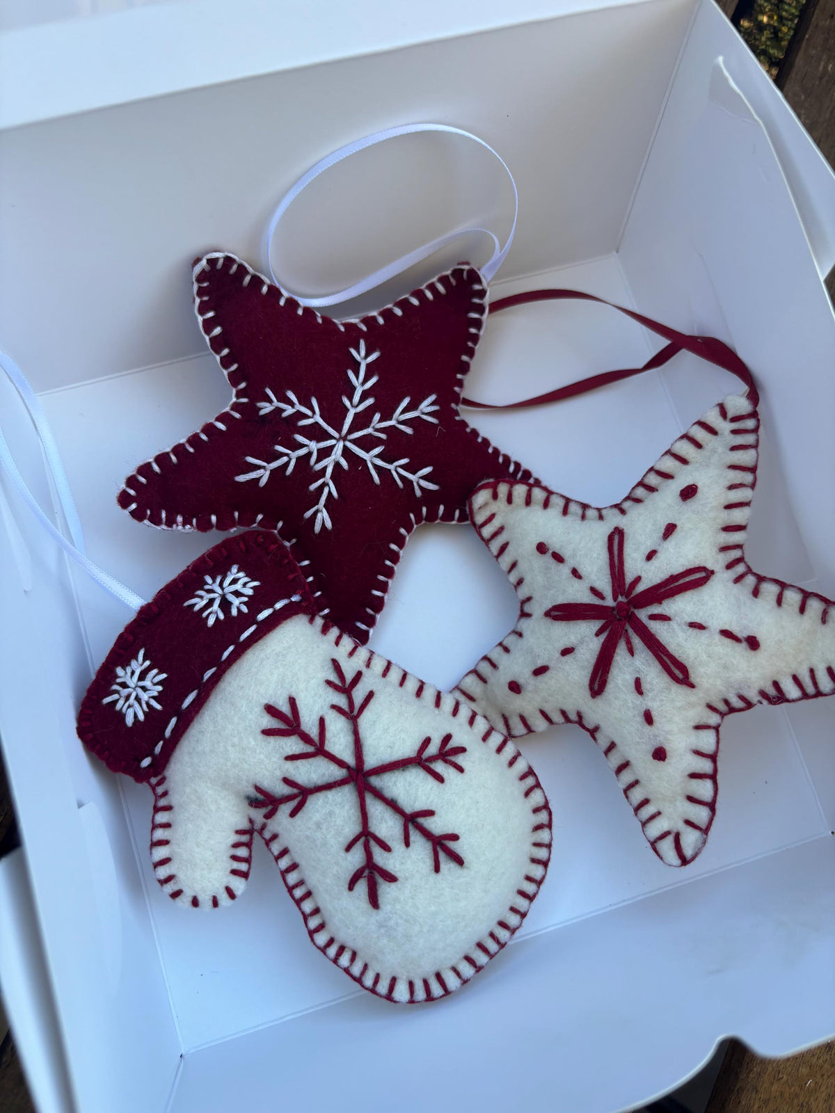 May include: A set of three Christmas ornaments in a white box. The ornaments include a burgundy star with a white snowflake, a white star with burgundy accents, and a white mitten with a burgundy cuff and snowflake design. Each ornament has a white ribbon for hanging.
