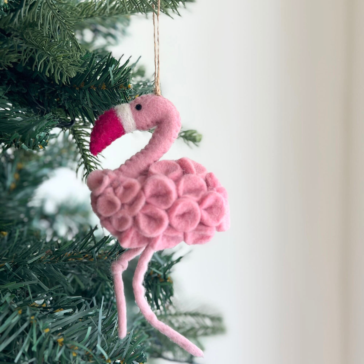 May include: Pink felt flamingo Christmas ornament with a white stripe on its beak. The ornament is hanging from a tree branch.