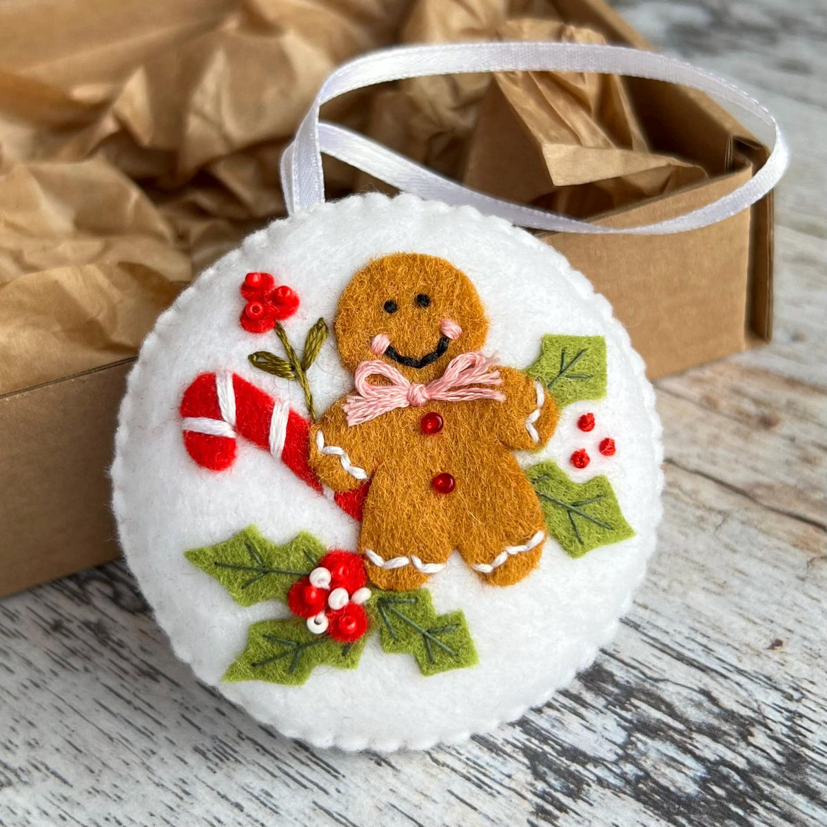 May include: A round, white felt Christmas ornament featuring a gingerbread man with a pink bow tie, a red and white candy cane, and green holly leaves with red berries. A white ribbon is attached for hanging.