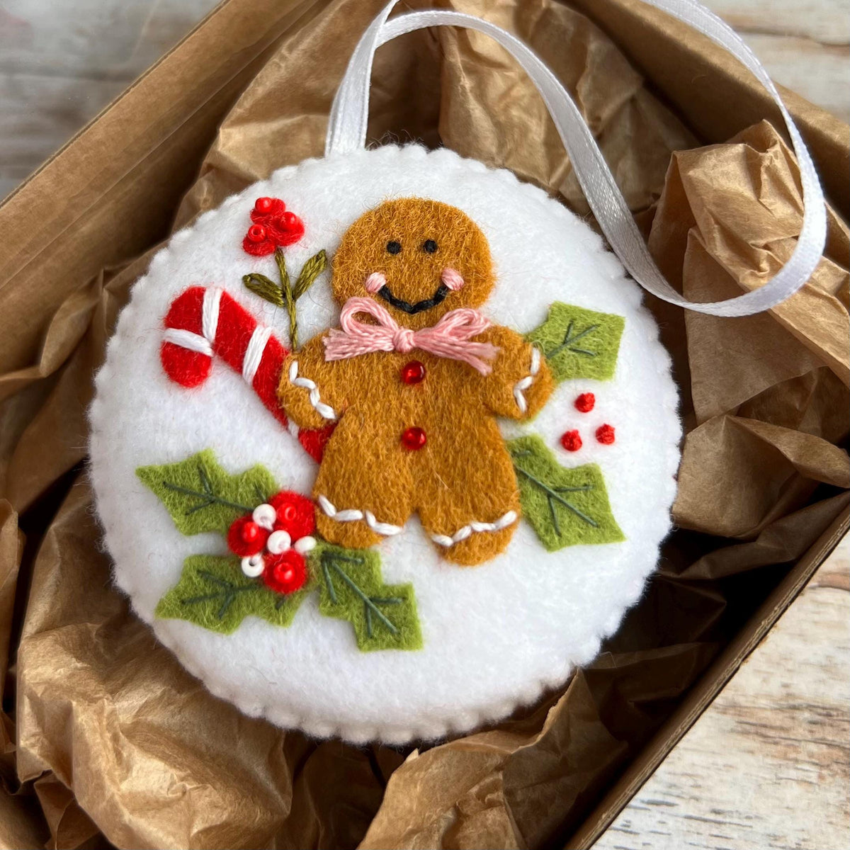 May include: A white felt Christmas ornament featuring a gingerbread man with a pink bow tie, a red and white candy cane, and holly leaves with red berries. The ornament has a white ribbon for hanging.