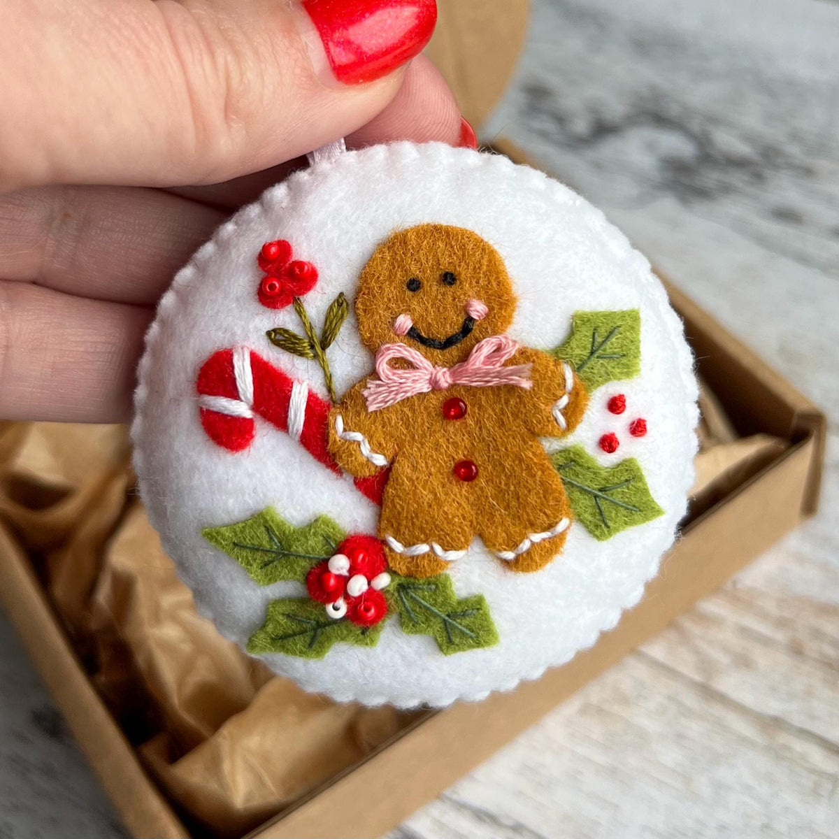 May include: A white felt Christmas ornament featuring a gingerbread man, candy cane, and holly. The gingerbread man has a pink bow tie and red buttons. The ornament is round and has a white ribbon for hanging. The ornament is inside a cardboard box.