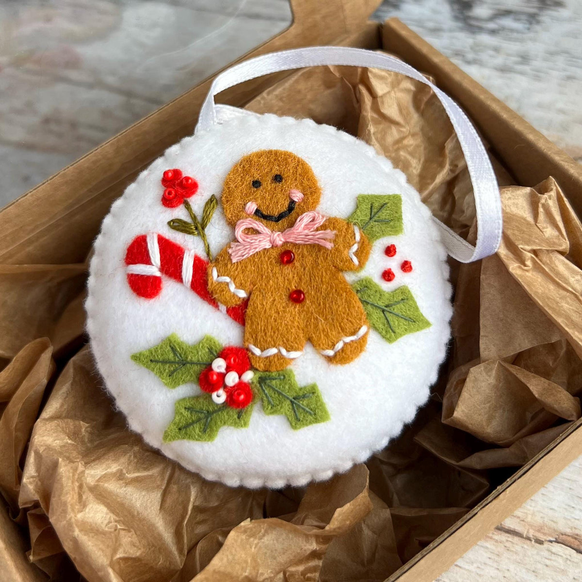 May include: A round, white Christmas ornament featuring a felt gingerbread man with a pink bow tie, a candy cane, and holly leaves with red berries. The ornament has a white ribbon for hanging and is presented in a brown box.