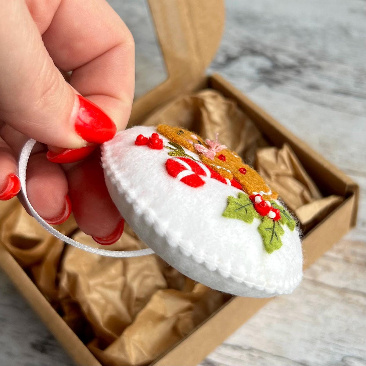 May include: A white felt Christmas ornament with a gingerbread man design. The ornament features red, green, and brown embroidered details, including candy canes and holly. It is held by a white string and is inside a cardboard box.