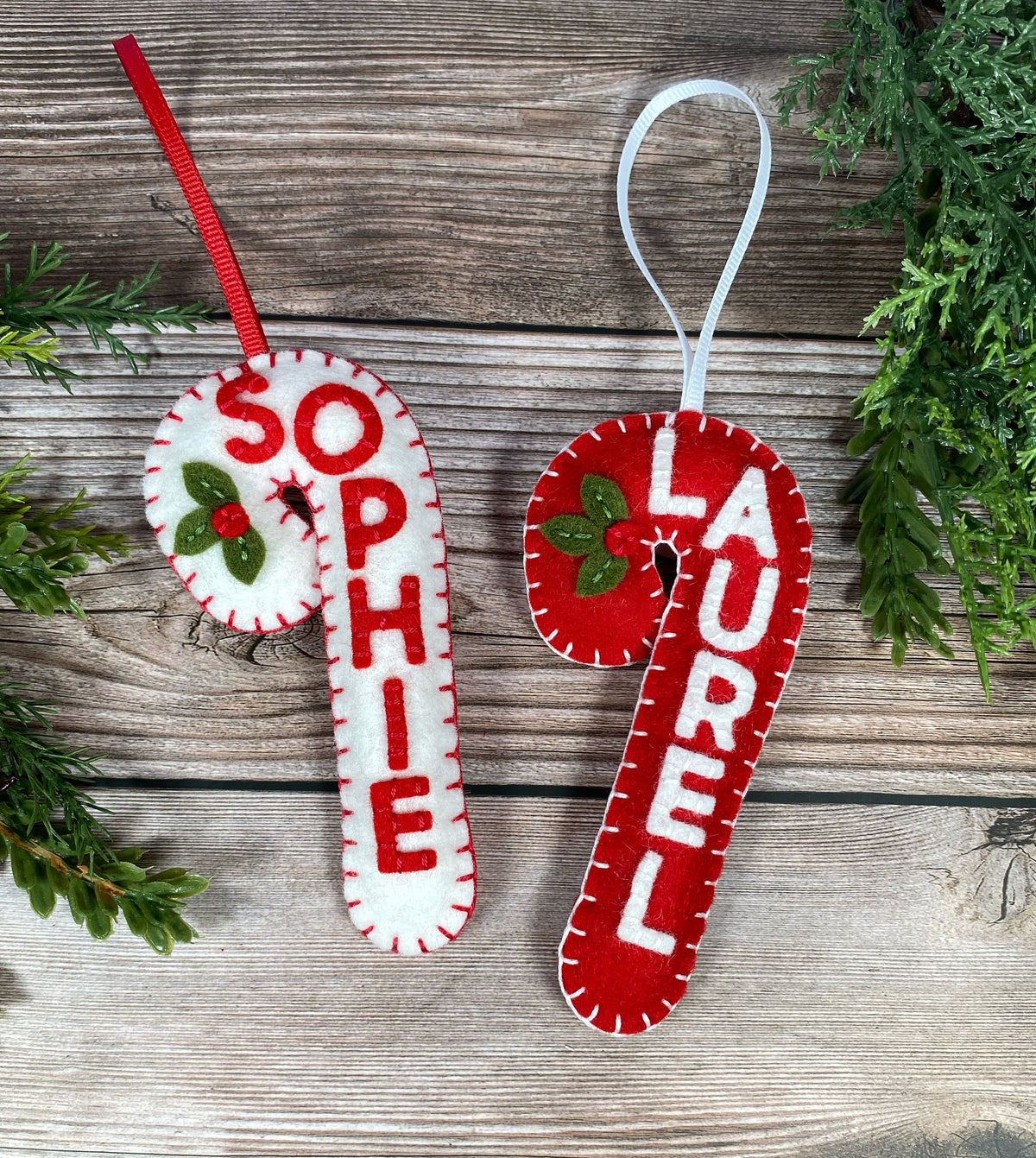 May include: Two felt candy cane ornaments with the names "Sophie" and "Laurel" stitched in white on a red and white background. The ornaments have green felt holly leaves and red berries.