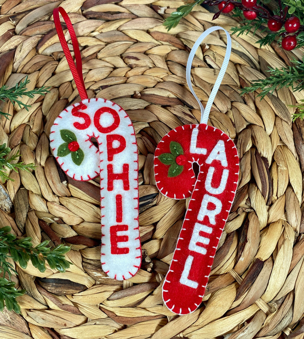 May include: Two felt candy cane ornaments with the names "Sophie" and "Laurel" embroidered on them. The ornaments are red and white with green holly leaves and red berries.