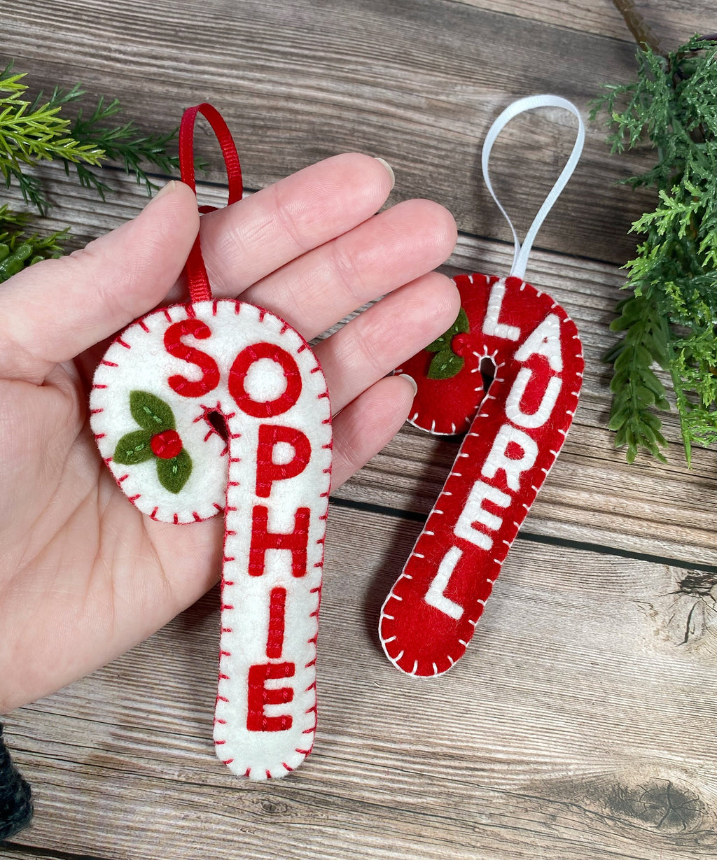 May include: Two felt candy cane ornaments with white felt and red felt. The candy canes are decorated with white stitching and the names "Sophie" and "Laurel" are written in white on the red felt.