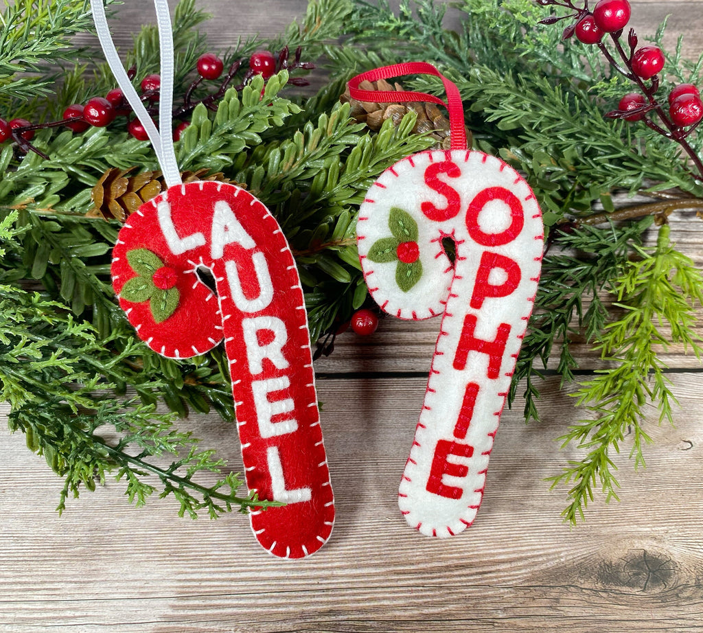 May include: Two felt candy cane ornaments with the names "Laurel" and "Sophie" embroidered on them. The ornaments are red and white with green holly accents.