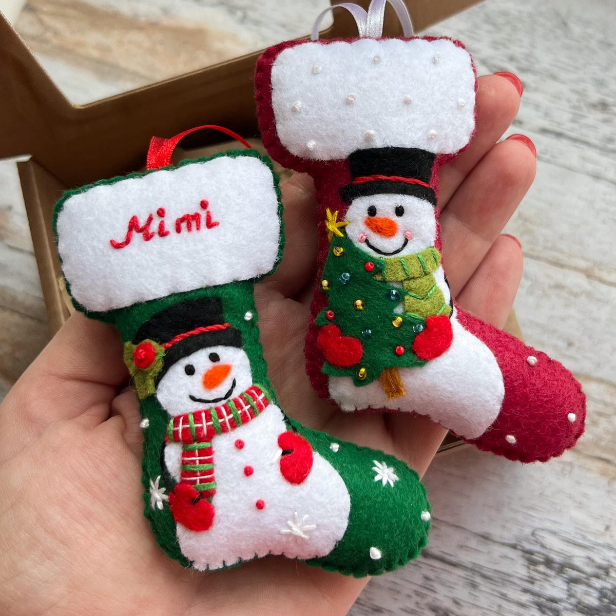 May include: Two felt Christmas stocking ornaments. One is green with a snowman wearing a red and green scarf and the name Mimi embroidered in red. The other is red with a snowman holding a green Christmas tree. Both have white cuffs.