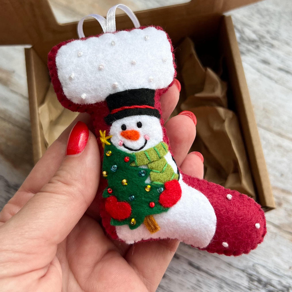 May include: A red felt Christmas stocking ornament featuring a white cuff with small white beads. The stocking displays an embroidered snowman wearing a black top hat and a green scarf, holding a green Christmas tree decorated with tiny beads.