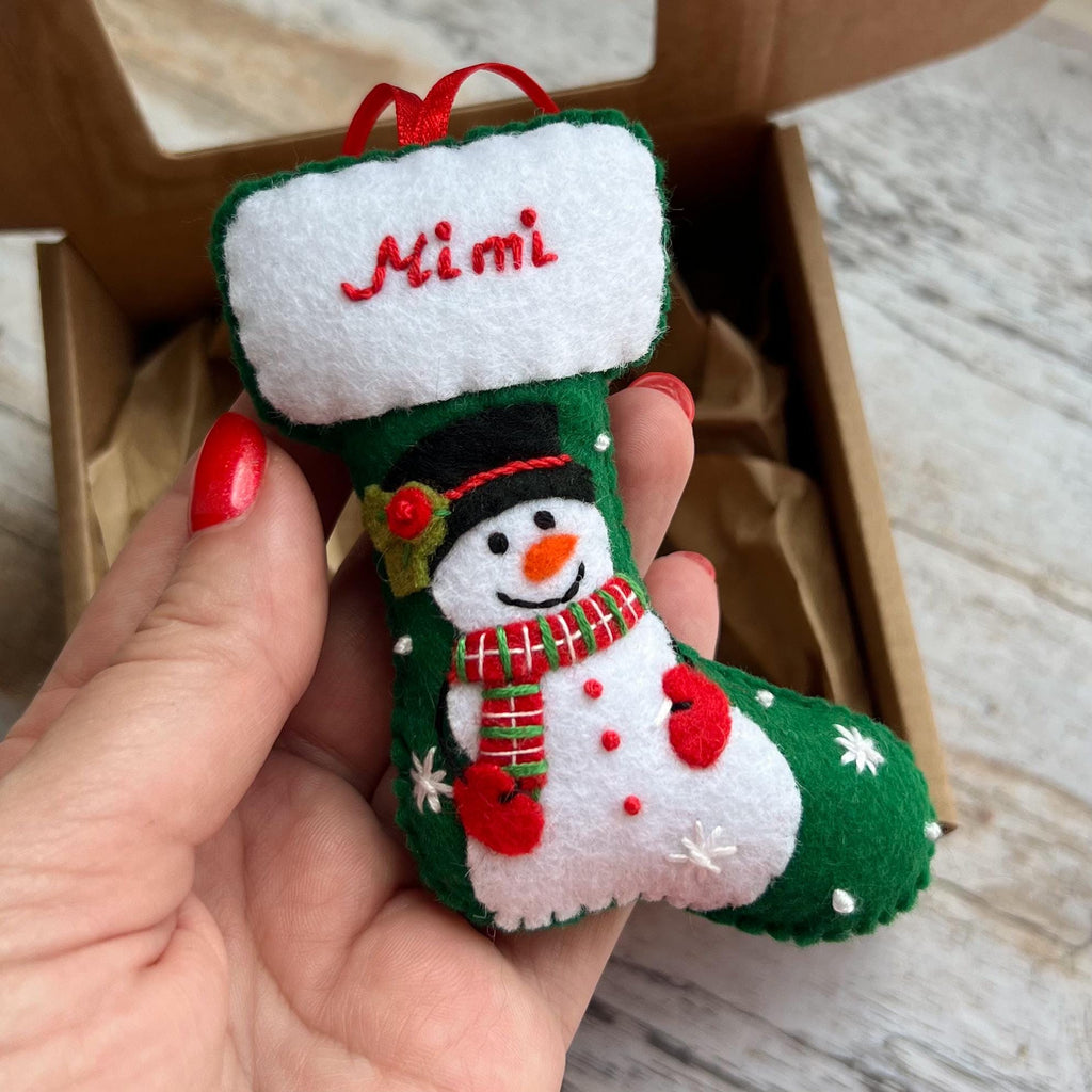 May include: A small felt Christmas stocking ornament in green and white. The cuff is white with the name Mimi embroidered in red. The body features a snowman with a black hat, orange nose, and a red and green striped scarf, accented by white snowflakes.