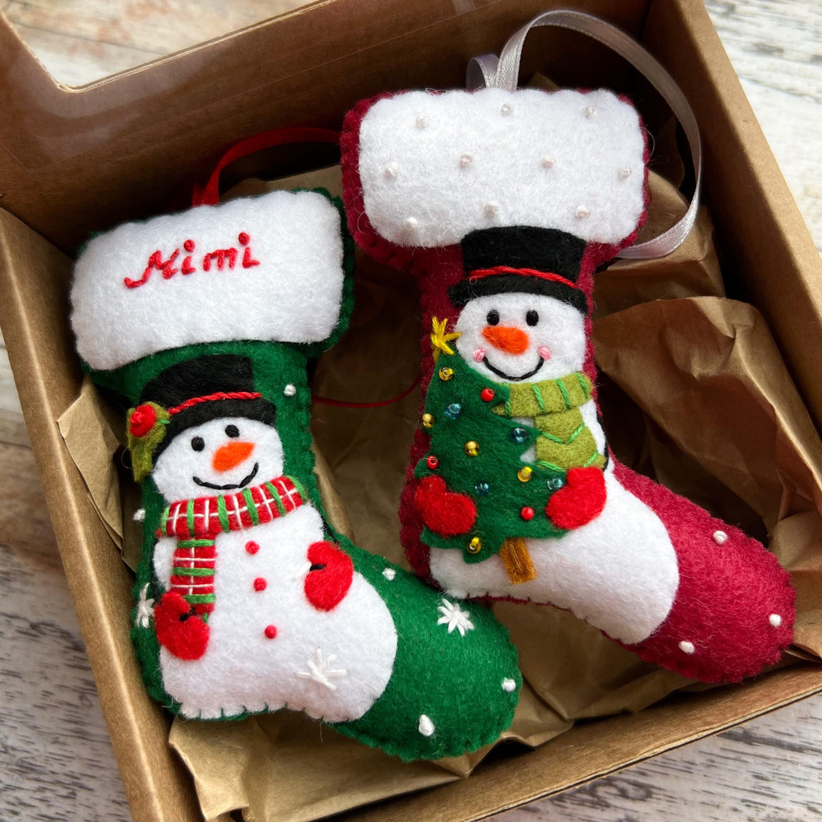 May include: Two felt Christmas stockings with snowman designs. The green stocking features the name Mimi embroidered in red, a snowman with a red and green scarf, and white snowflake accents. The red stocking features a snowman holding a green tree.