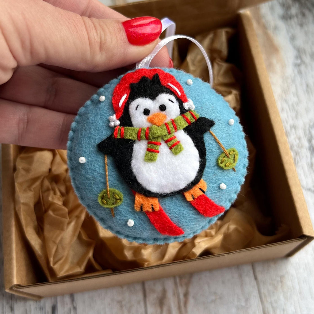 May include: A round felt ornament featuring a black and white penguin wearing a red hat and a green and red striped scarf. The penguin is skiing on a light blue background with small white beads representing snow and thin brown ski poles.