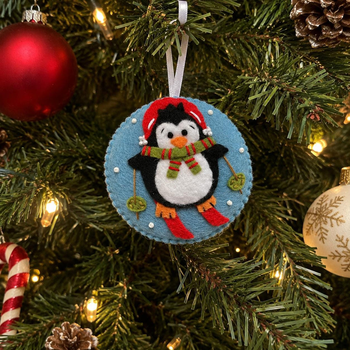 May include: A circular felt Christmas ornament featuring a black and white penguin wearing a red hat and a green and red striped scarf. The penguin is skiing on a light blue background with small white dots and holds two green ski poles.
