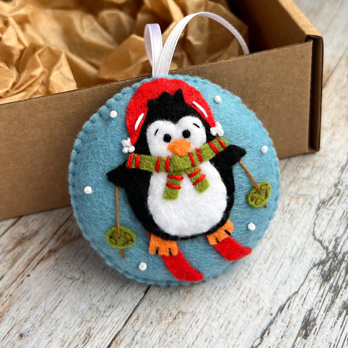 May include: A round felt ornament in light blue featuring a black and white penguin wearing a red hat, orange feet, and a green and red striped scarf. The penguin is skiing with red skis and poles, surrounded by small white embroidered dots.