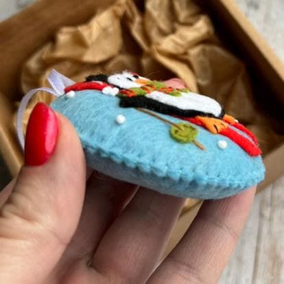 May include: A round, light blue felt ornament featuring a stitched penguin wearing a red and green scarf. The penguin is positioned on a light blue background with small white dots and a green circular accent. A white ribbon loop is attached to the top.