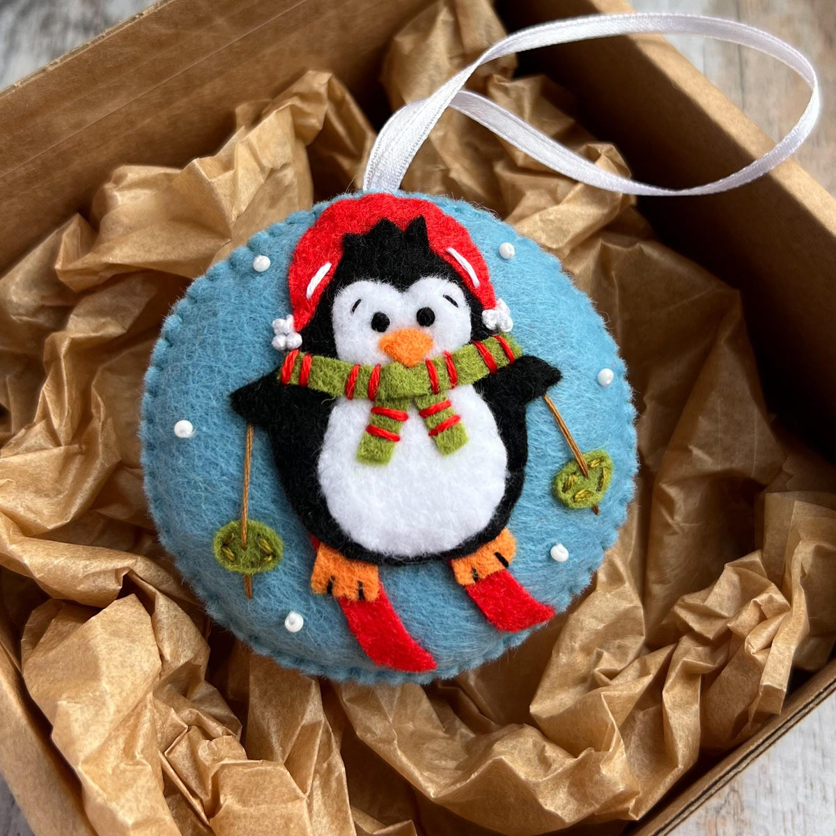 May include: A round felt ornament featuring a black and white penguin wearing a red hat and a green and red striped scarf. The penguin is skiing on a light blue background with small white dots representing snow and has a white ribbon loop.