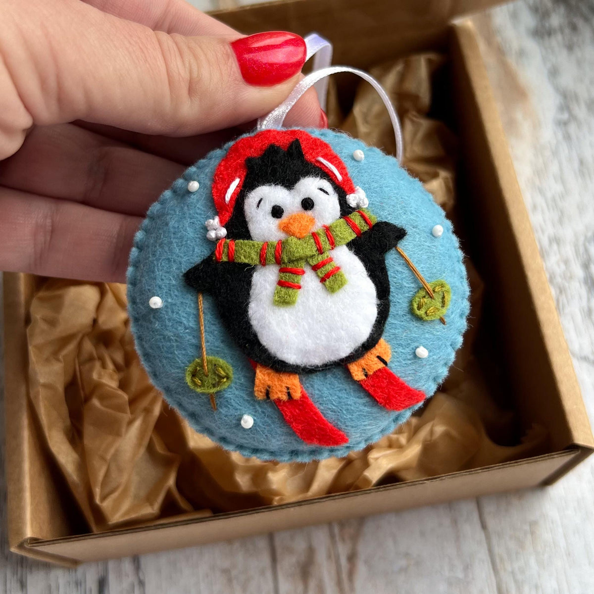 May include: A round felt ornament featuring a black and white penguin wearing a red hat and a green and red striped scarf. The penguin is skiing on a light blue background with small white beads representing snow and thin brown ski poles.