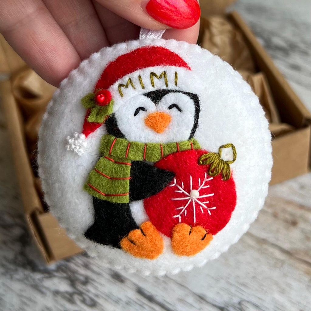 May include: A round, white Christmas ornament featuring a felt penguin wearing a red Santa hat with the name "MIMI" in gold letters. The penguin has a green scarf and holds a red ornament with a white snowflake design. The ornament has a white ribbon for hanging.