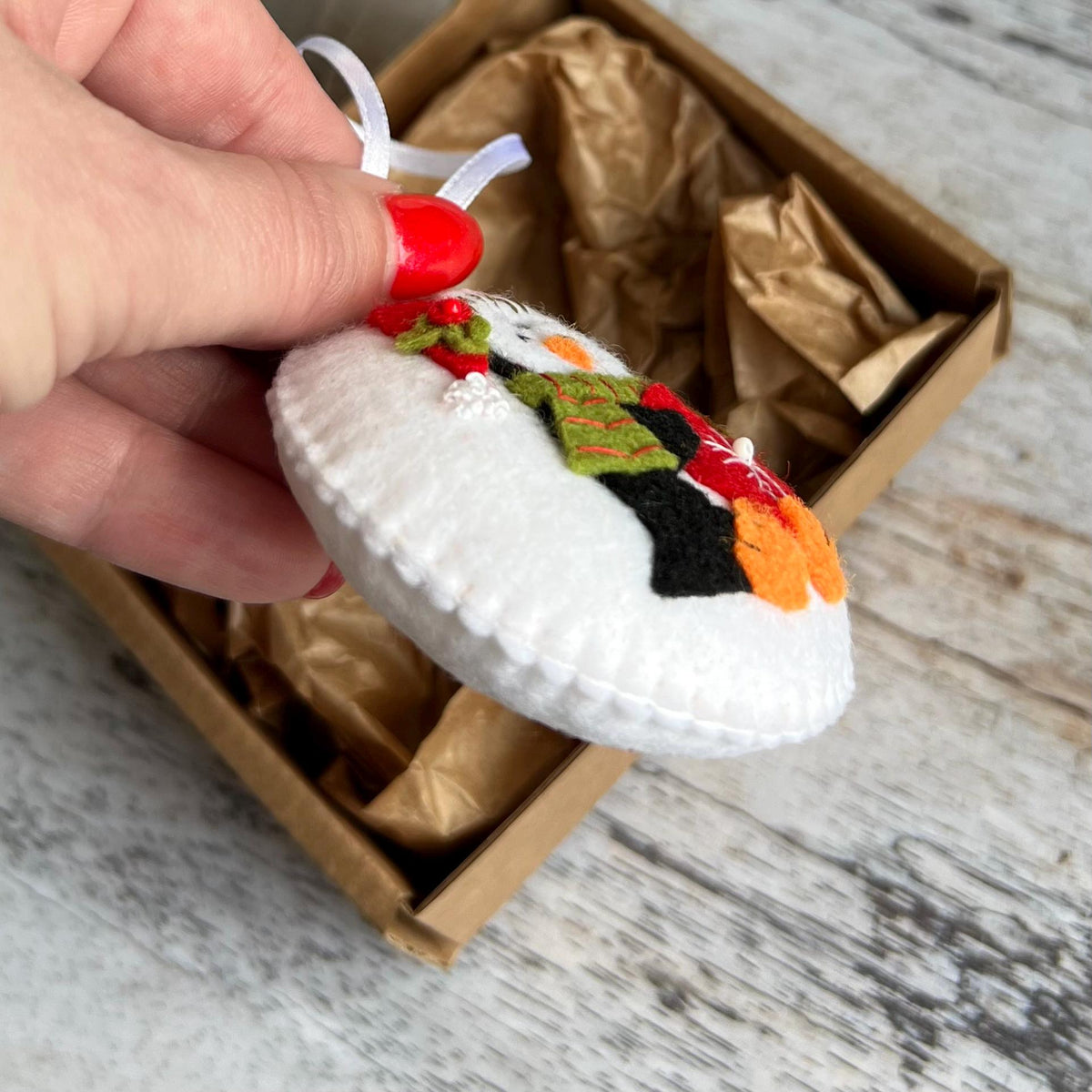 May include: A handmade felt snowman ornament with a white body, black top hat, and colorful scarf. The snowman has orange mittens and a red berry sprig. It is held by a white ribbon and presented in a brown box.