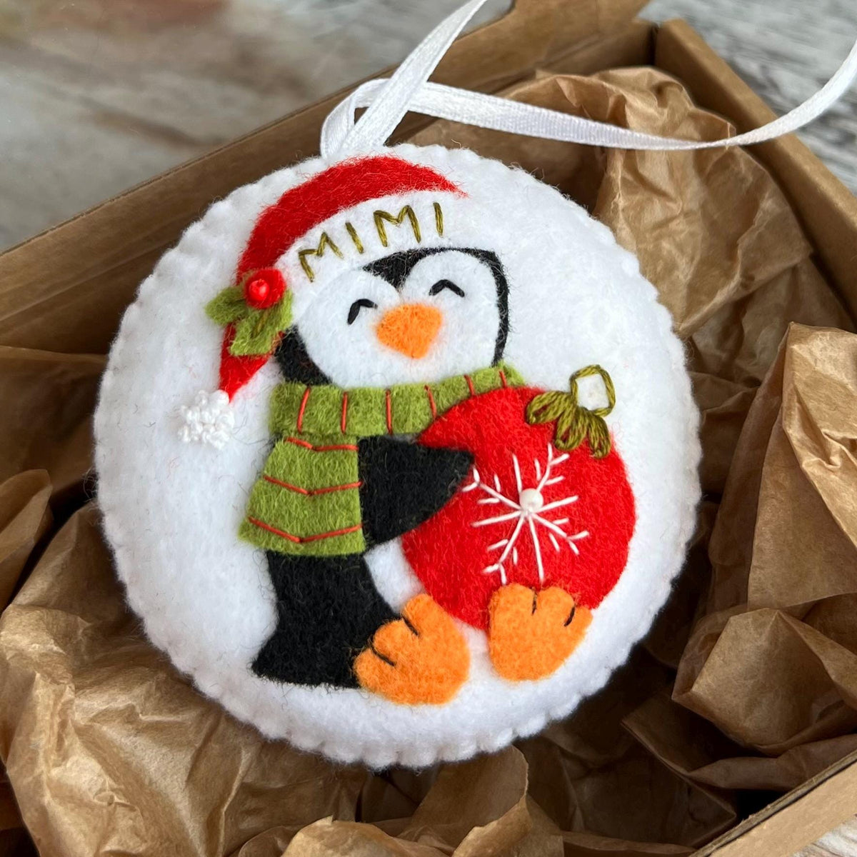 May include: A round, white felt Christmas ornament featuring a penguin wearing a red Santa hat with "MIMI" embroidered on it, a green scarf, and holding a red ornament with a white snowflake design. The ornament has a white ribbon for hanging.
